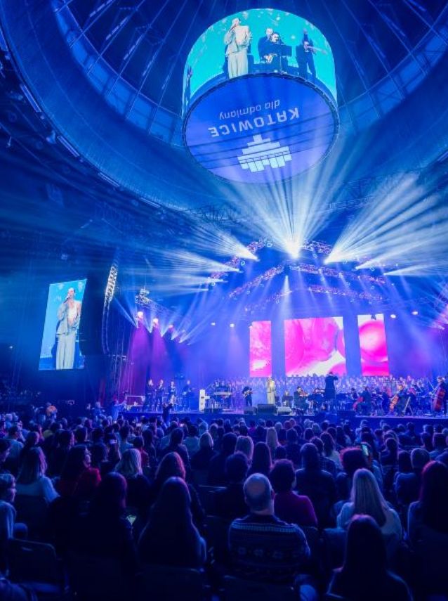 Liczna publiczność ogląda koncert na żywo na arenie z kolorowymi światłami, ekranami wyświetlającymi wykonawców oraz okrągłym ekranem nad sceną, na którym można zobaczyć szczegóły wydarzenia.