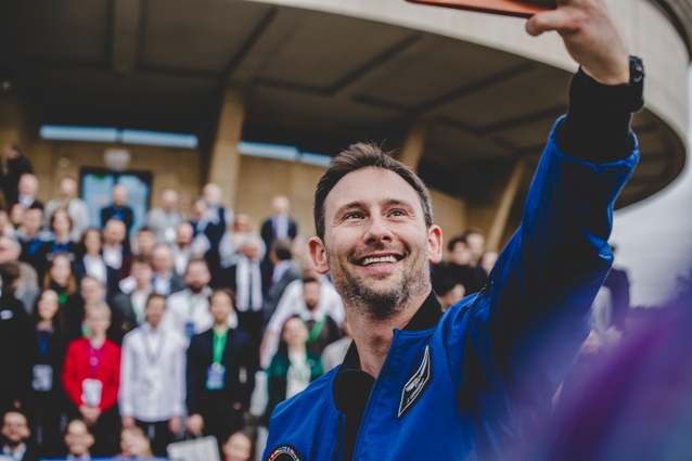 Uśmiechnięty mężczyzna w niebieskim kombinezonie astronauty robi sobie selfie przed dużą grupą ludzi stojących na schodach na zewnątrz nowoczesnego budynku. Tłum w tle jest lekko nieostry.