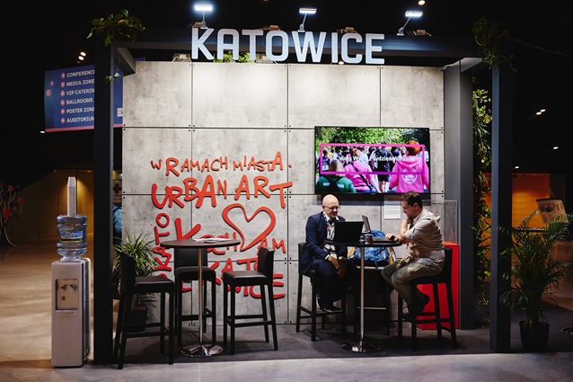 Dwóch mężczyzn siedzi przy wysokim stole pod stoiskiem z napisem KATOWICE, z muralem w stylu graffiti i ekranem pokazującym grupę osób w różu. W stoisku znajdują się rośliny, krzesła i dystrybutor wody w pobliżu.