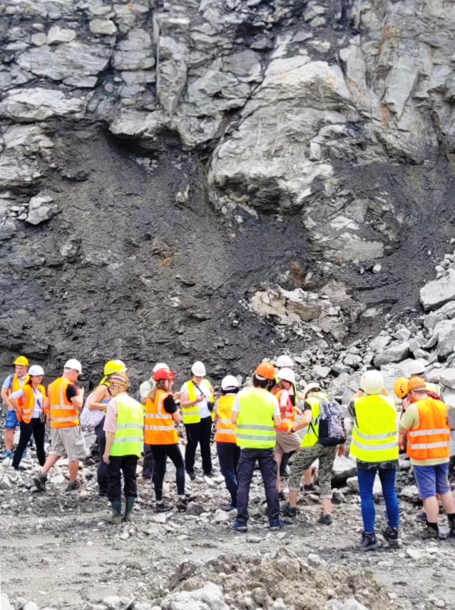 Grupa osób ubranych w kamizelki odblaskowe i kaski stoi na skalistym podłożu w pobliżu dużej ściany skalnej. Być może biorą udział w wycieczce z przewodnikiem lub wizycie edukacyjnej w kamieniołomie lub na placu budowy.