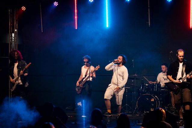 Zespół rockowy występuje na scenie w niebieskim i czerwonym świetle. Wokalista śpiewa namiętnie, dwóch gitarzystów, basista i perkusista grają za nimi, a publiczność ich obserwuje.