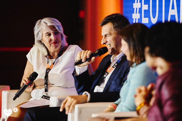 Cztery osoby siedzą na scenie podczas dyskusji panelowej. Jedna kobieta w bieli trzyma notatnik, podczas gdy mężczyzna obok niej mówi do mikrofonu. Dwóch innych panelistów jest częściowo widocznych na pierwszym planie.