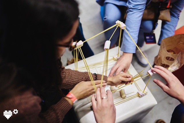 Trzy osoby używają surowego spaghetti i pianek, aby zbudować wysoką, geometryczną strukturę na stole, prawdopodobnie w ramach budowania zespołu lub aktywności twórczej. Ręce aktywnie układają patyczki spaghetti.