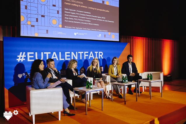 Sześciu panelistów siedzi na scenie z mikrofonami, dyskutując pod dużym napisem #EUTALENTFAIR i wyświetlanymi informacjami panelowymi. Na stołach znajdują się butelki i notatniki; scena jest oświetlona pomarańczowym światłem.
