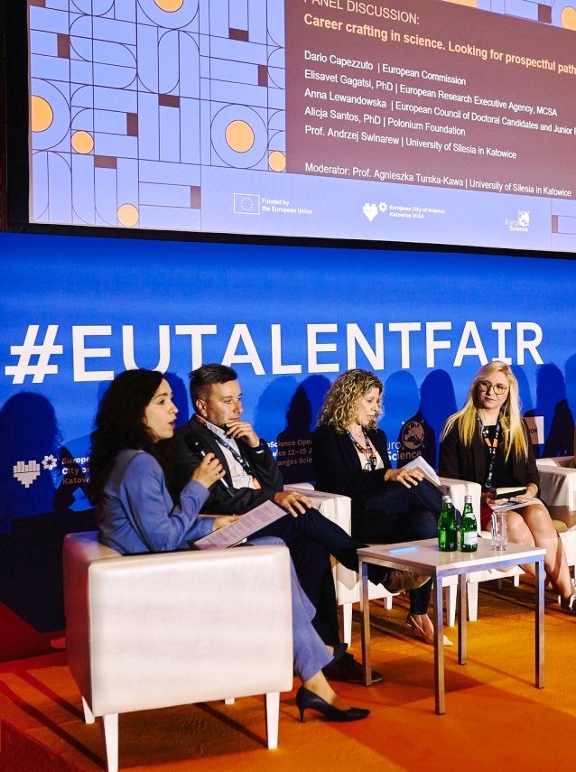 Pięciu panelistów siedzi na scenie z mikrofonami podczas dyskusji na wydarzeniu EUTalentFair. Za nimi znajduje się niebieskie tło z dużym białym napisem #EUTALENTFAIR. Na małym stoliku znajdują się butelki z wodą i papiery.
