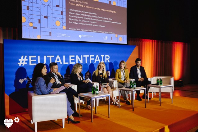 Sześciu panelistów siedzi na scenie z mikrofonami i dokumentami, dyskutując przed dużym niebiesko-pomarańczowym tłem z napisem #EUTALENTFAIR. Na stole znajdują się butelki wody i szklanki. Za nimi wyświetlany jest slajd z wydarzenia.