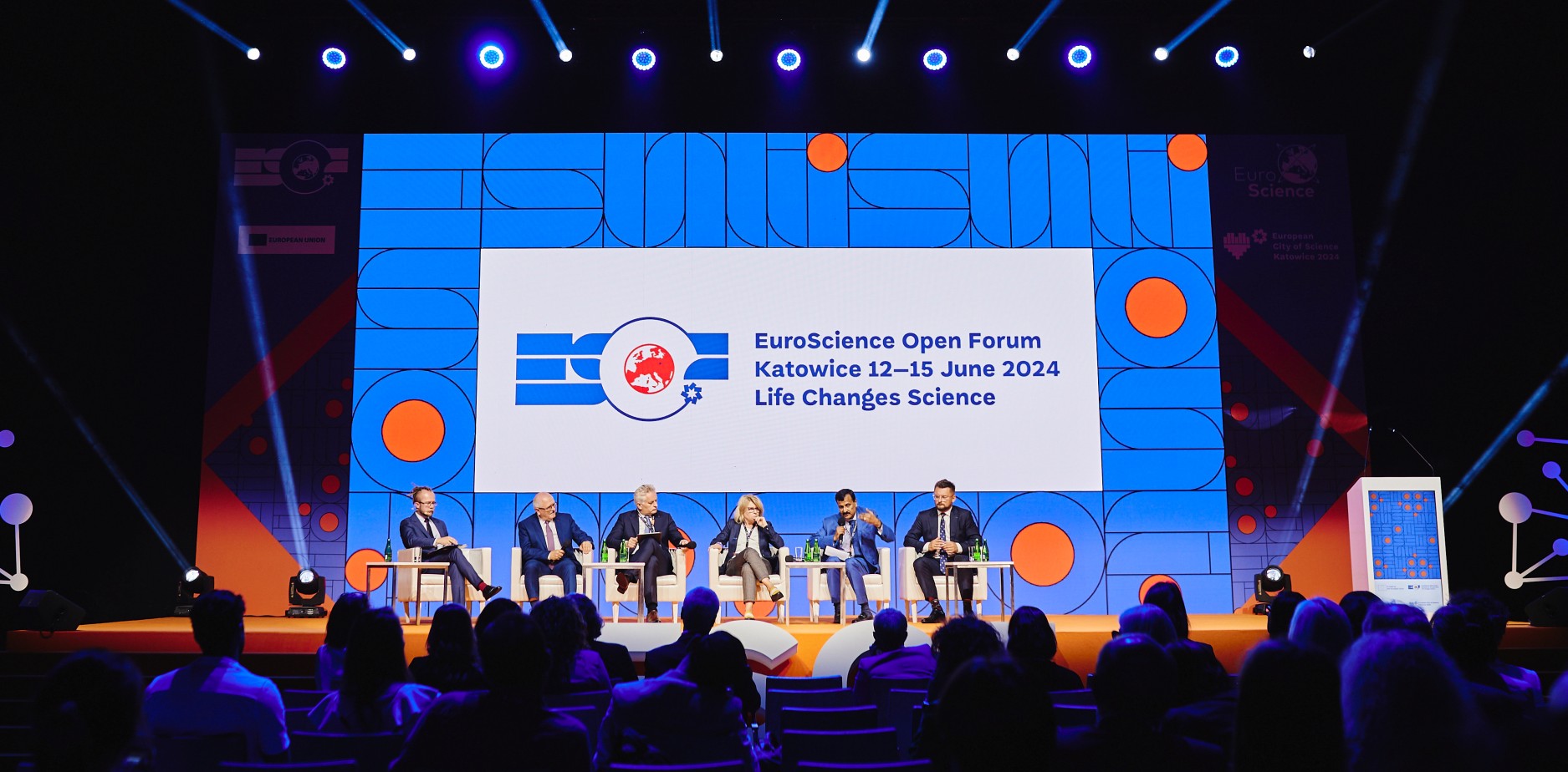 Sześcioosobowa grupa przemawia na scenie podczas EuroScience Open Forum w Katowicach. Za nimi znajduje się duży ekran, na którym wyświetlane są szczegóły wydarzenia, a z przodu siedzą członkowie widowni.