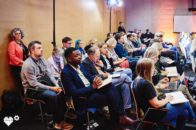 Zróżnicowana grupa osób siedzi i stoi w sali konferencyjnej, uważnie słuchając prezentacji. Niektórzy robią notatki na papierze lub laptopach; w pomieszczeniu są drewniane ściany, a w tle widoczny jest ekran projektora.