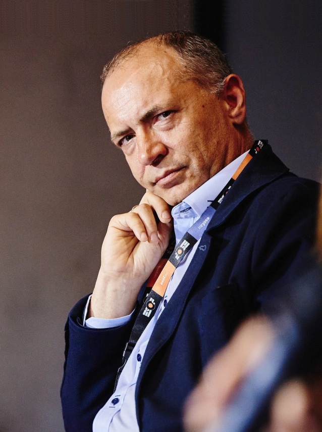 Mężczyzna w granatowej marynarce i ze smyczką siedzi zamyślony, opierając rękę na brodzie i patrząc prosto w kamerę na tle delikatnie oświetlonego tła.