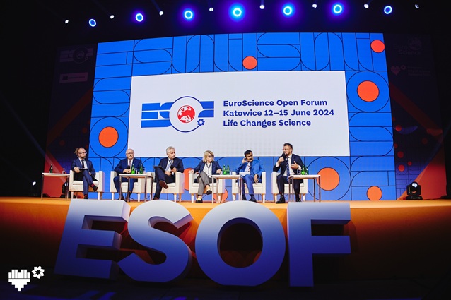 Sześć osób siedzi na scenie przed dużym ekranem wyświetlającym logo EuroScience Open Forum i szczegóły wydarzenia: „Katowice 12–15 czerwca 2024, Życie zmienia naukę”. Scena i tło są jasno oświetlone.