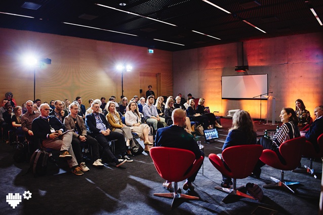 Grupa ludzi siedzi w rzędach naprzeciwko panelu czterech mówców na czerwonych krzesłach podczas konferencji w pomieszczeniu. Duży ekran i jasne światła są ustawione z przodu nowoczesnego, przestronnego pomieszczenia.