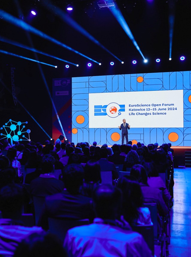 Prelegent stoi na scenie podczas EuroScience Open Forum 2024 w Katowicach, przemawiając do publiczności. Na scenie wyświetlane są szczegóły wydarzenia za pomocą kolorowych grafik i jasnoniebieskiego oświetlenia.