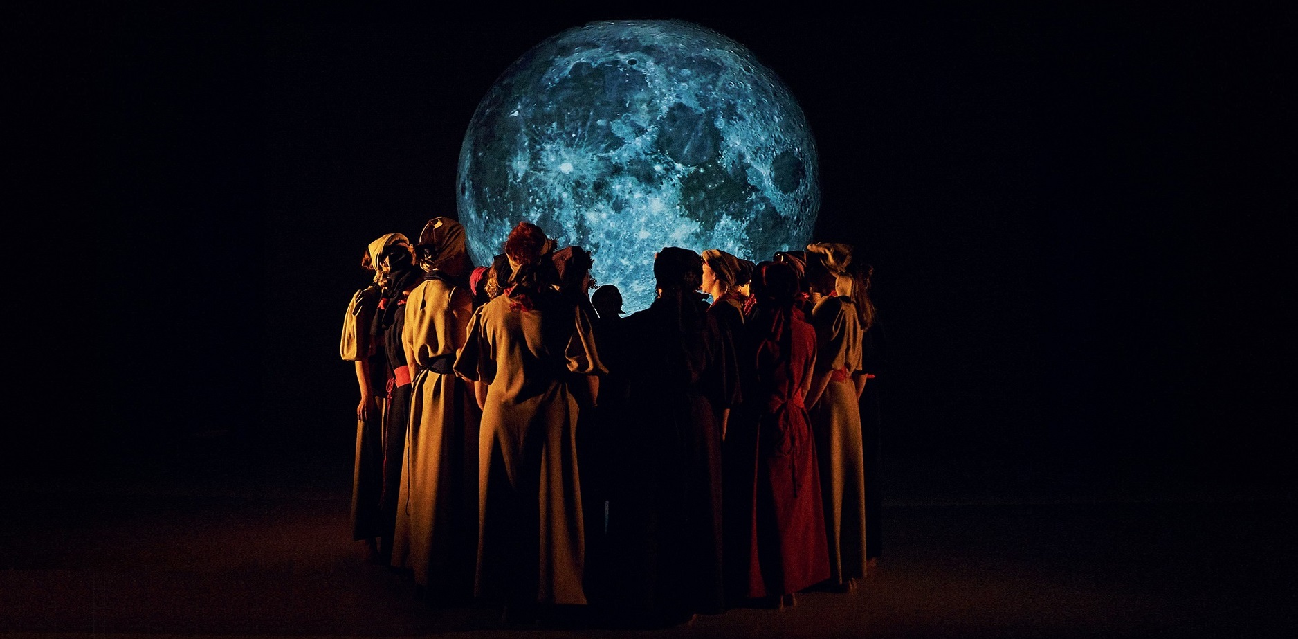 Grupa ludzi ubranych w długie szaty stoi w kręgu, zwrócona twarzą do dużego, oświetlonego księżyca na ciemnej scenie. Scena jest dramatyczna, księżyc rzuca niebieską poświatę na grupę.