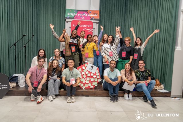 Zróżnicowana grupa młodych dorosłych pozuje i uśmiecha się entuzjastycznie na tle zielonej kurtyny i znaku EU Talenton, a niektórzy z nich trzymają symbole pokoju i notatki umieszczone na tablicy w środku.