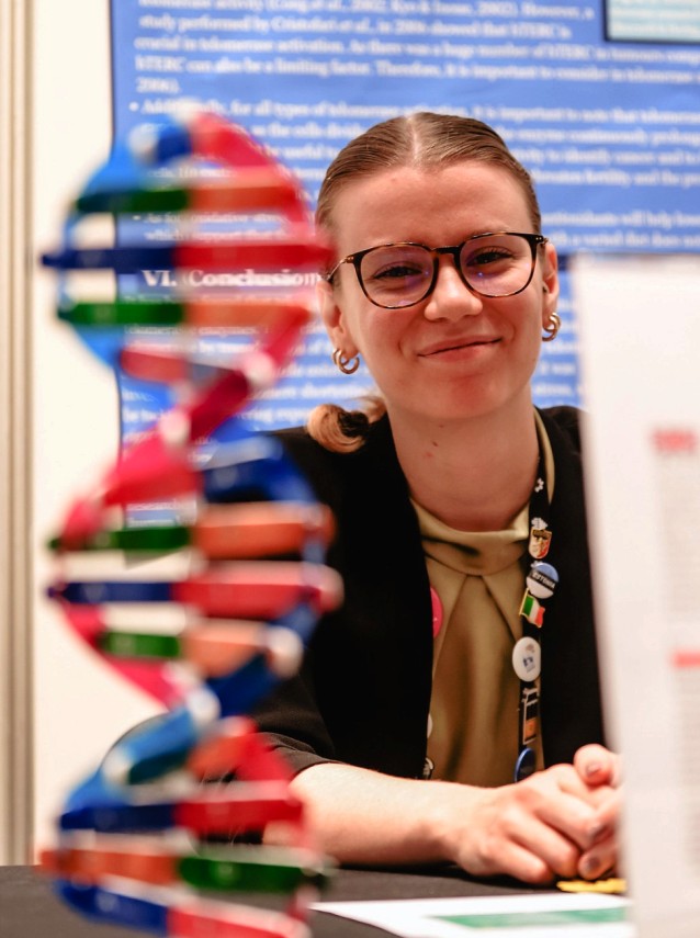 Osoba w okularach i czarnej marynarce z przypinkami uśmiecha się, stojąc za kolorowym modelem podwójnej helisy DNA, w tle widnieje niebiesko-biały plakat badawczy.