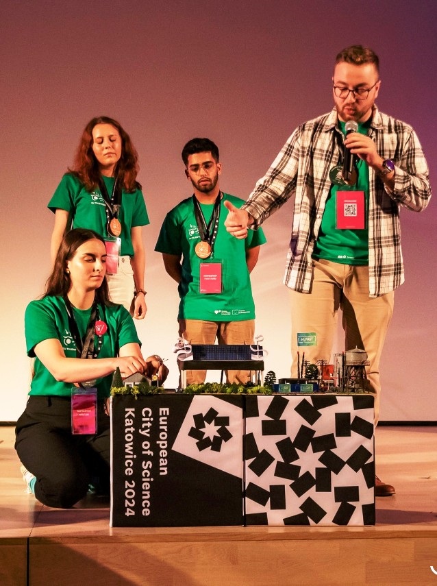 Cztery osoby w zielonych koszulkach stoją na scenie i prezentują projekt naukowy, którego model leży na stole podpisanym Europejskie Miasto Nauki Katowice 2024. Jedna osoba mówi do mikrofonu, podczas gdy pozostali uważnie słuchają.