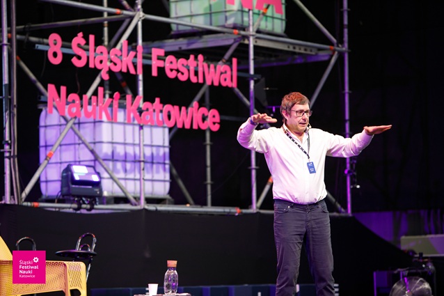 Mężczyzna w białej koszuli i okularach przemawia na scenie z rozłożonymi ramionami podczas 8. Śląskiego Festiwalu Nauki w Katowicach. Za nim wyświetlana jest nazwa festiwalu dużymi różowymi literami.