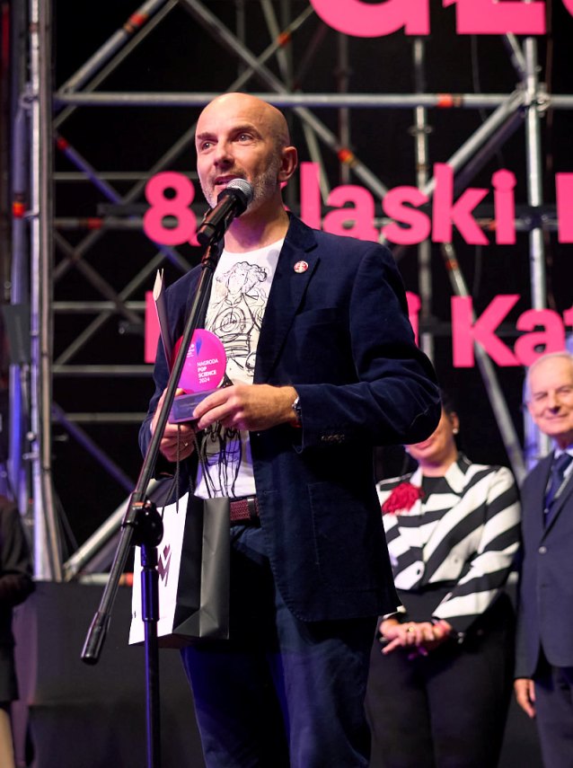 Mężczyzna w ciemnym blezerze stoi na scenie, trzymając nagrodę i mówiąc do mikrofonu, z torbą prezentową w ręku. Kilka osób stoi za nim, a na tle rusztowania widoczny jest duży różowy tekst.