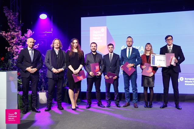 Grupa ośmiu osób w formalnych strojach stoi na scenie trzymając certyfikaty, z dużym ekranem i różowym oświetleniem w tle. Jedna osoba trzyma ogromny czek.