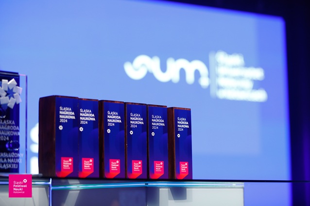 Pięć prostokątnych drewnianych nagród z napisem ŚLĄSKA NAGRODA NAUKOWA 2024 ustawiono na stole z niebieskim tłem, na którym widnieje rozmyte logo wydarzenia i tekst.
