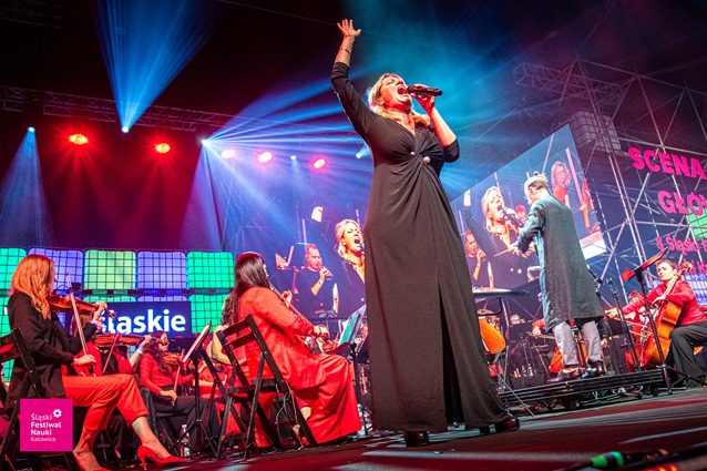 Piosenkarka w czarnej sukience występuje na scenie z orkiestrą ubraną na czerwono. Trzyma mikrofon i podnosi jedną rękę, a duże ekrany za nią wyświetlają jej wizerunek, a kolorowe światła sceniczne oświetlają scenę.