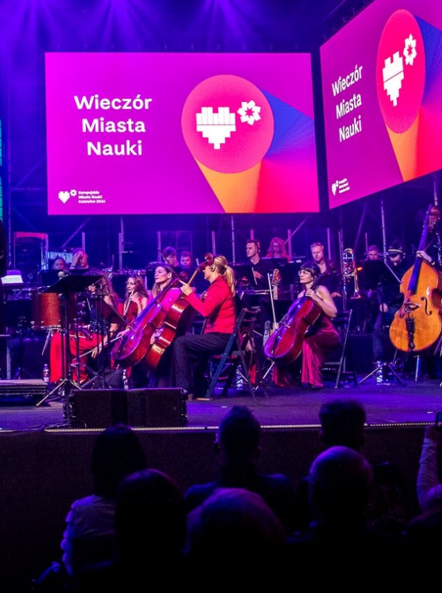 Orkiestra symfoniczna występuje na scenie pod fioletowym oświetleniem, a muzycy grają na instrumentach strunowych i dętych. Za nimi na dwóch dużych ekranach wyświetla się „Wieczór Miasta Nauki” z grafiką serca i kwiatów.