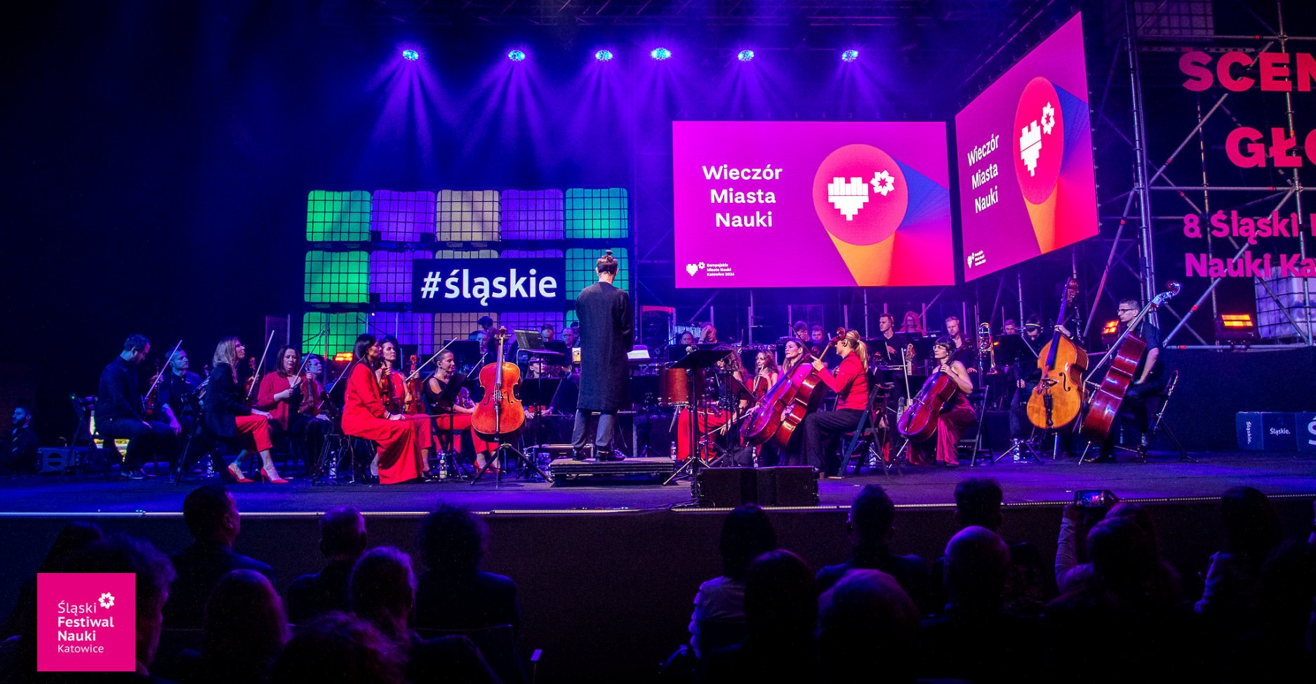 Orkiestra występuje na scenie pod fioletowym i niebieskim oświetleniem na Śląskim Festiwalu Nauki w Katowicach, a muzycy są ubrani na czerwono i czarno. Na ekranach wyświetla się „Wieczór Miasta Nauki” i logo wydarzenia. Na pierwszym planie widać publiczność.