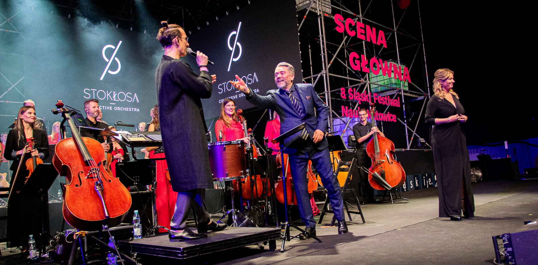 Orkiestra gra na żywo na jasno oświetlonej scenie. Muzycy grają na instrumentach strunowych, podczas gdy dwóch prezenterów w formalnych strojach przemawia i ściska dłonie. Kobieta w czarnej sukience stoi z boku. W tle widoczne są napisy w języku polskim.