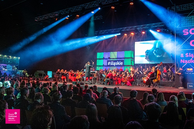 Duża orkiestra występuje na jasno oświetlonej scenie przed siedzącą publicznością na festiwalu naukowym. Ekrany za muzykami wyświetlają wizualizacje i tekst, a kolorowe światła oświetlają scenę.