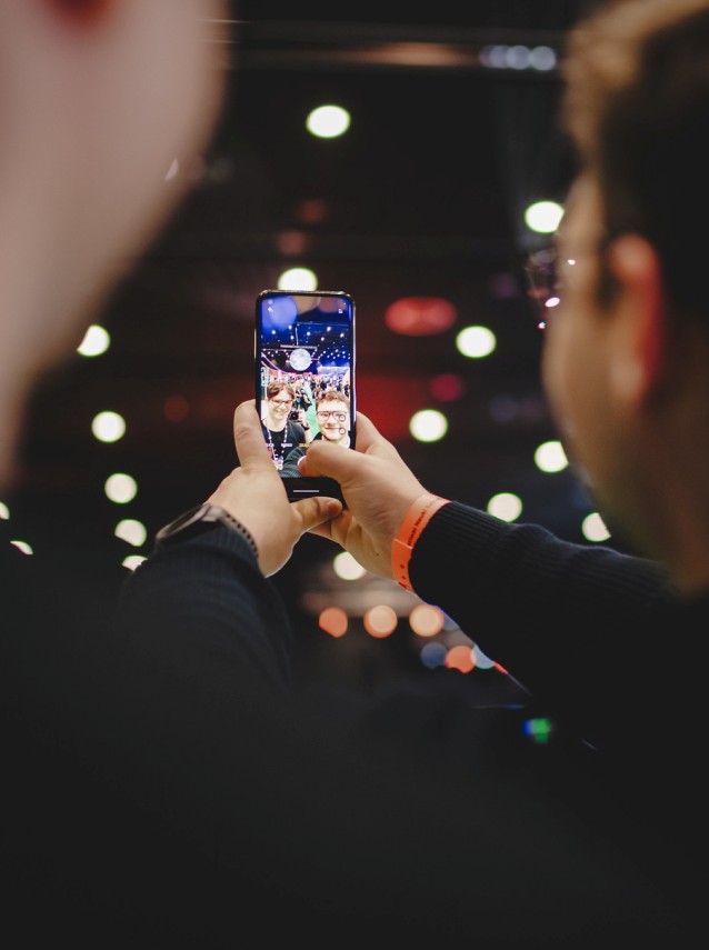 Dwie osoby robią sobie selfie smartfonem na imprezie z jasnymi światłami bokeh w tle. Jedna osoba ma na sobie pomarańczową opaskę. Na ekranie telefonu widać ich uśmiechnięte twarze.