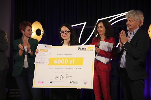 Kobieta trzyma w ręku duży czek na kwotę 6000 zł podczas konkursu naukowego FameLab, otoczona trzema osobami klaszczącymi na scenie za ciemnymi kurtynami i z widocznymi w tle tablicami informacyjnymi o wydarzeniu.