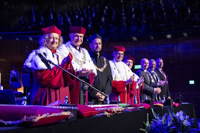 Grupa rektorów w ozdobnych togach i czapkach akademickich stoi w rzędzie na scenie podczas oficjalnej ceremonii, uśmiecha się i klaszcze, a w słabo oświetlonym tle widać publiczność.