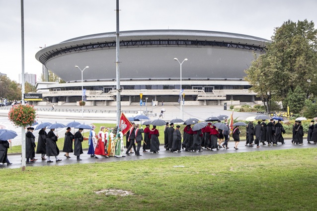 Przemarsz ludzi w akademickich togach i niosących parasole przechodzi obok Spodka w Katowicach w pochmurny dzień. Niektórzy ludzie niosą sztandary, a na pierwszym planie widać drzewa i trawę.