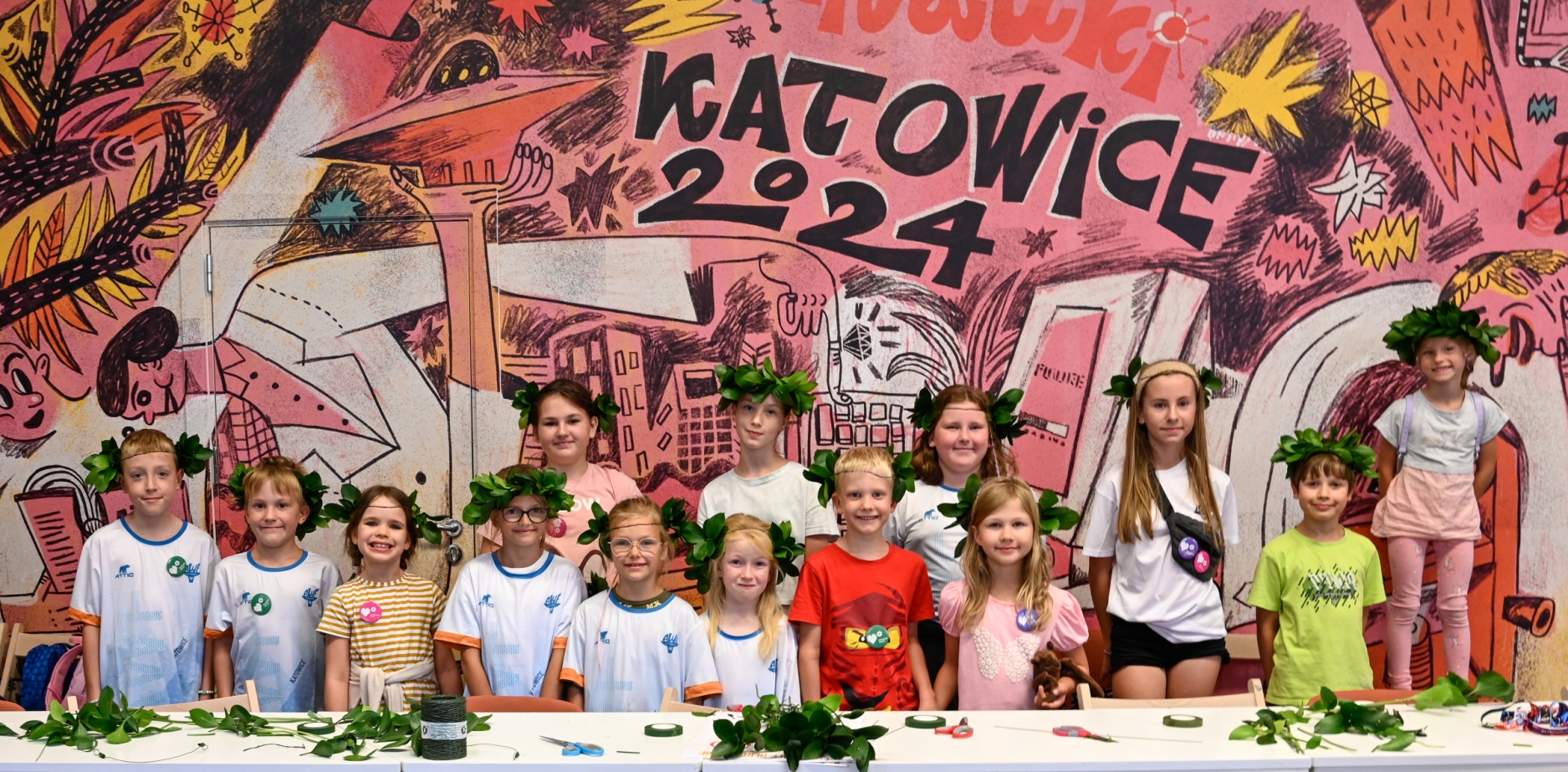 Grupa dzieci i dwóch młodych dorosłych w koronach z liści pozują i uśmiechają się przed kolorowym muralem z napisem „Katowice 2024”. Stoją za stołem ozdobionym zielonymi liśćmi.