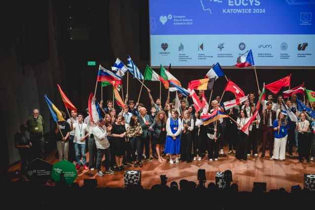 Duża grupa młodych ludzi stoi na scenie, trzymając flagi różnych państw podczas wydarzenia naukowego, a za nimi na ekranie wyświetla się EUCYS Katowice 2024 i loga kilku sponsorów.