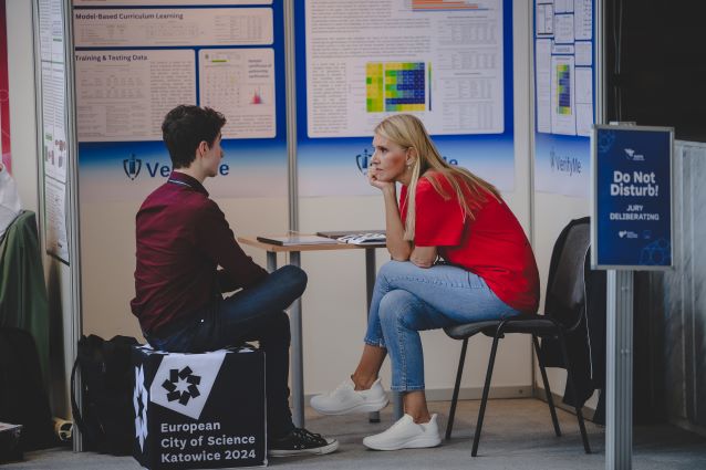 Kobieta w czerwonej koszuli i dżinsach siedzi naprzeciwko mężczyzny w ciemnym ubraniu przy małym stoliku, rozmawiając na wydarzeniu naukowym. W tle widoczne są plakaty i znak Do Not Disturb!.