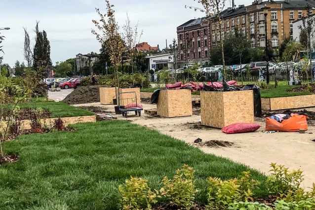 Miejski park w budowie z zieloną trawą, młodymi drzewami, drewnianymi skrzynkami na sadzonki, workami z ziemią i wózkiem na utwardzonej ścieżce. W tle widoczne są budynki mieszkalne i zaparkowane samochody.