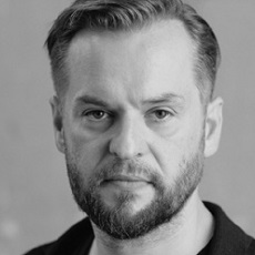 Czarno-biały portret mężczyzny z krótkimi włosami i brodą, patrzącego bezpośrednio w kamerę z neutralnym wyrazem twarzy. Ma na sobie ciemną kurtkę na jasnej koszuli.