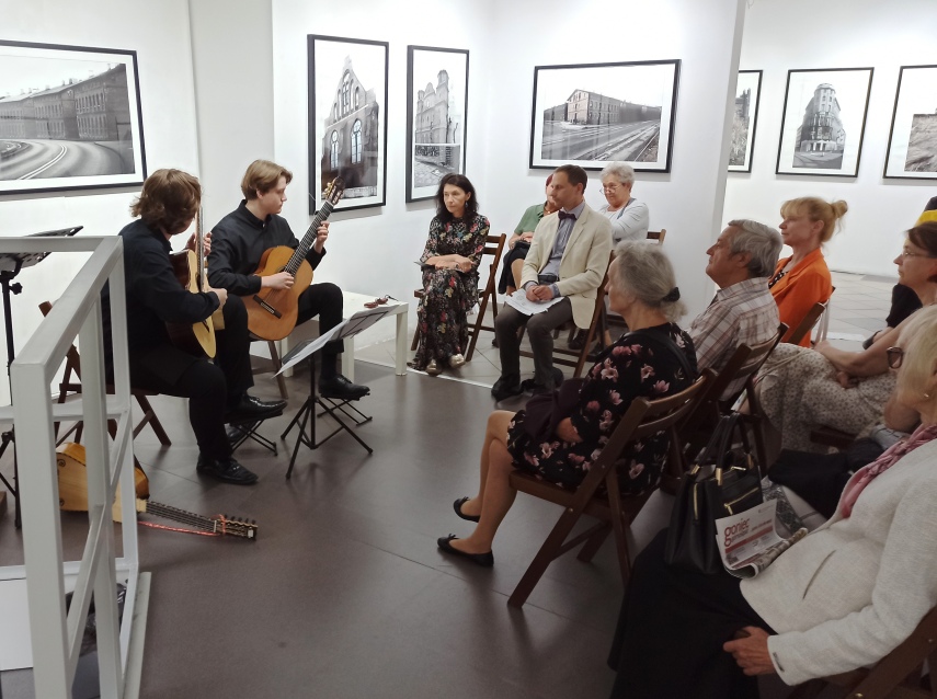 Dwóch muzyków gra na gitarach klasycznych dla siedzącej publiczności w galerii sztuki. Na ścianach wokół nich wiszą czarno-białe fotografie budynków. Publiczność słucha uważnie.