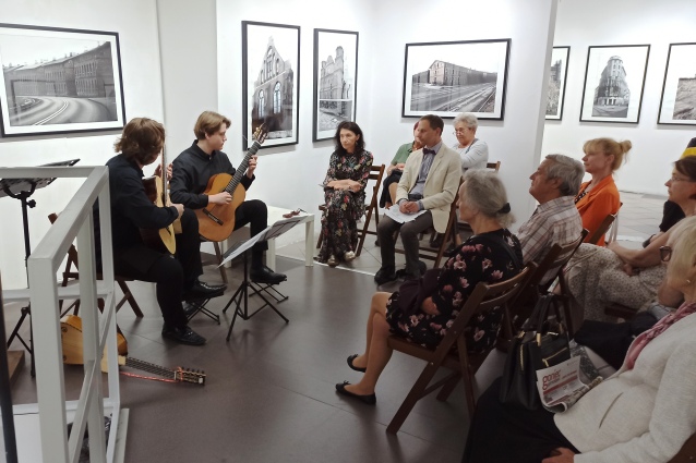 Dwóch gitarzystów gra dla małej siedzącej publiczności w galerii sztuki. Na białych ścianach wiszą czarno-białe fotografie budynków. Słuchacze wydają się być uważni, otoczeni nowoczesną, minimalistyczną scenerią.