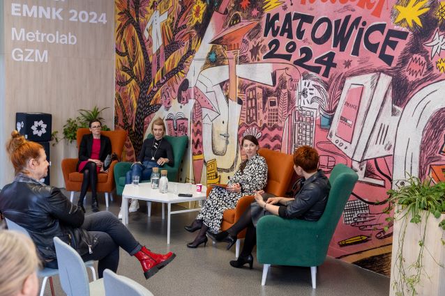 Pięć kobiet siedzi na krzesłach i dyskutuje przed kolorowym muralem z napisem „Katowice 2024”. Trzy kobiety siedzą po jednej stronie, a dwie po drugiej, między nimi jest stół i drinki.