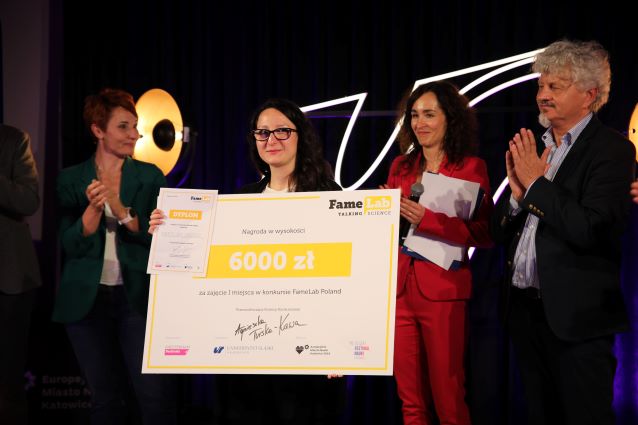 Kobieta trzymająca duży czek na 6000 zł stoi na scenie z czterema innymi osobami, w tym dwiema kobietami klaszczącymi i jedną trzymającą mikrofon, podczas ceremonii wręczania nagród. W tle znajduje się logo FameLab.
