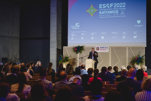 Prelegent stoi na podium, zwracając się do publiczności na ESOF 2022 EuroScience Open Forum w Katowicach. Scena ma duży ekran z informacjami o wydarzeniu i ozdobnymi roślinami po obu stronach.