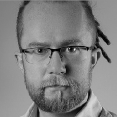 Czarno-biały portret osoby w okularach, z krótką brodą i wąsami. Jej włosy są częściowo ogolone, z kilkoma dredami po jednej stronie. Ma poważny wyraz twarzy i nosi koszulę z kołnierzykiem.