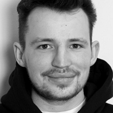 Młody mężczyzna z krótkimi ciemnymi włosami i zarostem uśmiecha się do kamery. Ma na sobie ciemną bluzę z kapturem i jest sfotografowany na jasnym tle.