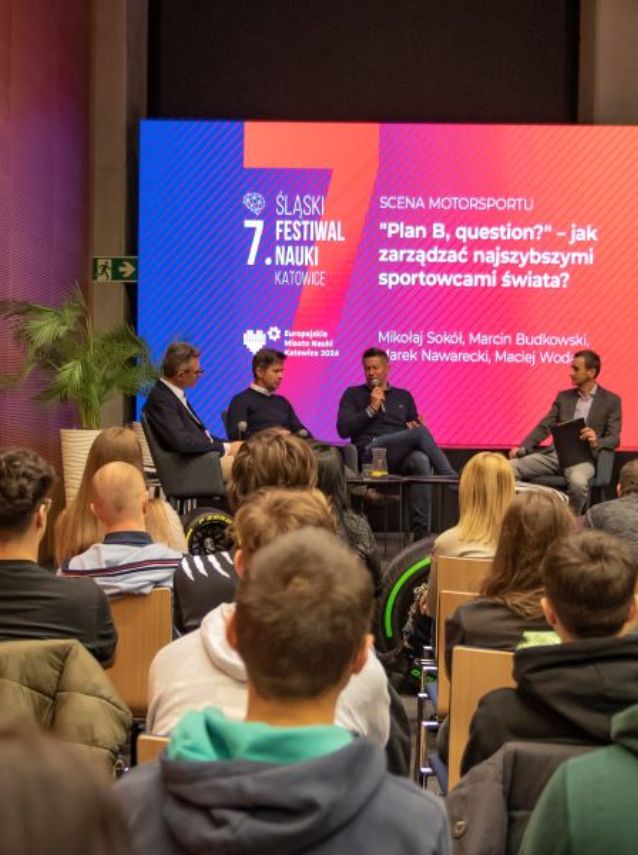 Czterech mówców zasiada na scenie i dyskutuje o zarządzaniu sportem motorowym podczas 7. Śląskiego Festiwalu Nauki w Katowicach. Publiczność przysłuchuje się wypowiedziom, a za nimi na dużym ekranie wyświetlany jest temat panelu oraz nazwiska uczestników.