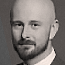 Portret w skali szarości przedstawiający łysego mężczyznę z brodą, ubranego w garnitur i krawat, stojącego zwróconego twarzą do aparatu z neutralnym wyrazem twarzy.