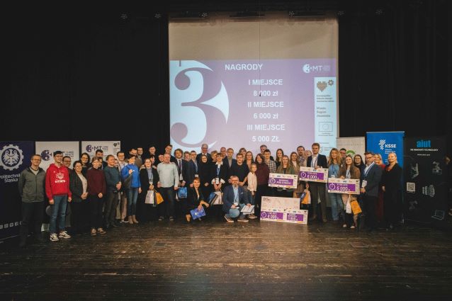 Duża grupa ludzi stoi na scenie, trzymając nagrody i torby z prezentami, pozuje do zdjęcia przed ekranem projektora, na którym wyświetlane są kwoty nagród i informacje o wydarzeniu w języku polskim.