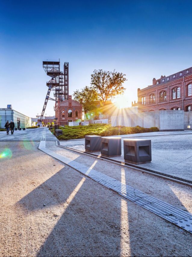 Nowoczesny miejski plac z betonowymi ławkami, drzewem i ceglanymi budynkami. Wpadające promienie słońca rzucają długie cienie. W tle widać wieżę przemysłową pod czystym, błękitnym niebem. W oddali spaceruje kilka osób.