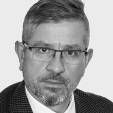 Mężczyzna z krótkimi, starannie uczesanymi włosami, w okularach, z przystrzyżoną brodą i wąsami, ubrany w kraciastą marynarkę, białą koszulę i krawat, patrzący bezpośrednio w kamerę z neutralnym wyrazem twarzy.